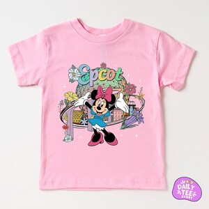 A pink t-shirt with a Minnie Mouse character and various colorful elements, such as flowers and a globe, printed on it.