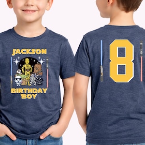 May include: A navy blue t-shirt featuring a Star Wars theme. The front displays the name "Jackson" above a graphic of characters including C-3PO, Chewbacca, and Darth Vader, with the text "BIRTHDAY BOY". The back shows a large yellow number "8" with lightsabers.