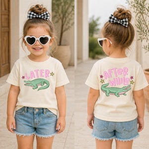 May include: A cream-coloured t-shirt featuring a green alligator graphic and the word "LATER" in pink on the front. The back of the shirt says "AFTER WHILE" with a matching alligator graphic. The child is wearing denim shorts.