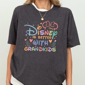 A person wearing a gray t-shirt with a colorful Disney-themed design and text that says "Disney is better with Grandkids".