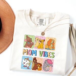 Op de afbeelding: Crèmekleurig T-shirt met kleurrijke cartoonafbeeldingen van Disney-personages. Het shirt heeft de woorden "MOM VIBES" in een speels lettertype. Een gouden ketting is over het shirt gedrapeerd.