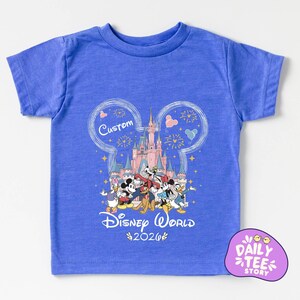 A blue t-shirt with a Disney World-themed graphic design, including Mickey Mouse, Donald Duck, and other Disney characters, along with a castle and fireworks. The text "Custom Disney World" and "Disney World 2026" is also visible.