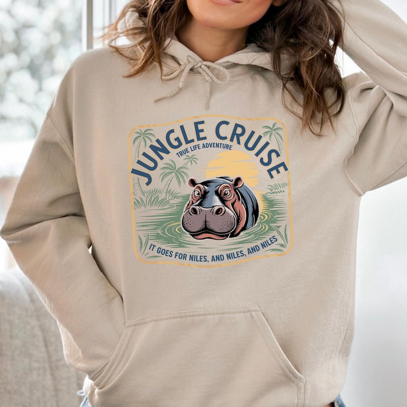 A person wearing a beige hoodie with a graphic design featuring a hippopotamus and palm trees, along with the text "JUNGLE CRUISE" and "IT GOES FOR MILES, NILES, AND MILES".