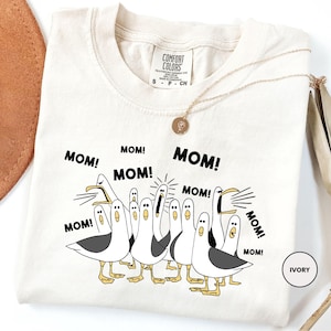 A white t-shirt with a graphic design of a group of seagulls and the text "MOM! MOM! MOM!" printed on it.