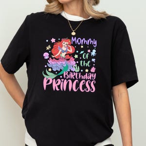 A person wearing a black t-shirt with a colorful graphic design featuring a mermaid character and text that says "Mommy of the Birthday Princess".