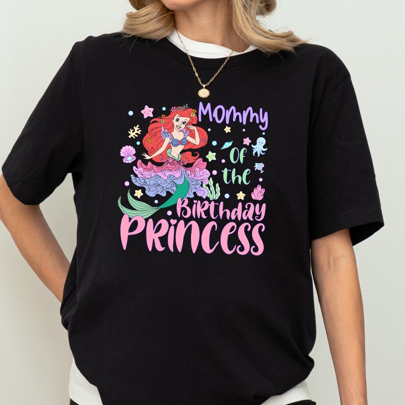 A person wearing a black t-shirt with a colorful graphic design featuring a mermaid character and text that says "Mommy of the Birthday Princess".