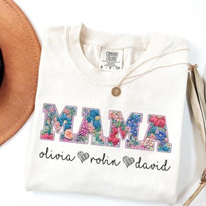 Op de afbeelding: Crèmekleurig T-shirt met het woord "MAMA" in bloemrijke letters. Onder het woord staan de namen "Olivia", "Rolin" en "David", elk met een hartsymbool. Een gouden ketting en een bruine hoed zijn ook zichtbaar.