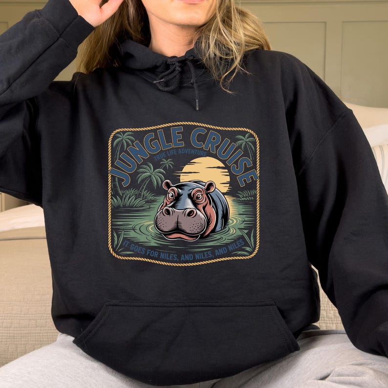 A person wearing a black hoodie with a graphic design featuring a hippopotamus and the text "JUNGLE CRUISE" printed on it.