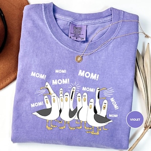 a purple t-shirt with a graphic design of a group of cartoon seagulls, each with a surprised expression, accompanied by the text "MOM! MOM! MOM! MOM!" in a playful and humorous manner.