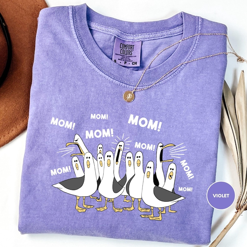 a purple t-shirt with a graphic design of a group of cartoon seagulls, each with a surprised expression, accompanied by the text "MOM! MOM! MOM! MOM!" in a playful and humorous manner.