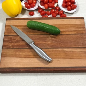 May include: A wooden cutting board with a light brown and dark brown striped pattern. A silver knife and a green cucumber are on the board.