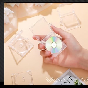 May include: Several clear plastic CD cases, some open and some closed, are arranged on a light-coloured surface. One case contains a reflective mini CD. The cases are square with a hinged lid, designed to protect the discs.