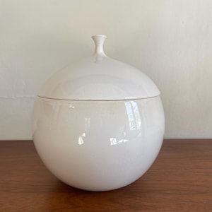 Pode incluir: Um pote branco de cerâmica com tampa. O corpo esférico tem um acabamento brilhante, e a tampa apresenta um pequeno botão arredondado. O pote está sobre uma superfície de madeira. Design simples e elegante.