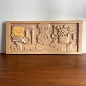 Könnte beinhalten: Wandkunst aus Holz mit Küchenmotiv, geschnitzt mit einem Koch und Küchenutensilien. Das Relief umfasst eine Kochmütze, einen Löffel, ein Messer und einen Topf. Das Kunstwerk ist aus hellem Holz gefertigt und hat eine rechteckige Form.