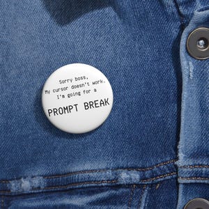 以下が含まれることがあります： 「Sorry boss, My cursor doesn't work. I'm going for a PROMPT BREAK」と黒い文字で書かれた白いボタンバッジ。青いデニムジャケットにピンが留められています。