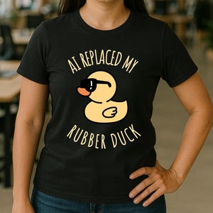 Peut inclure: T-shirt noir avec l'inscription "AI REPLACED MY RUBBER DUCK" et un canard de dessin animé portant des lunettes de soleil. Le canard est jaune avec un bec orange. Le t-shirt est à manches courtes.