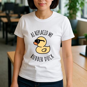 May include: A woman in a white t-shirt with the text "AI REPLACED MY RUBBER DUCK". The shirt has a cartoon yellow rubber duck wearing black sunglasses. The setting is an office, with desks and plants visible.