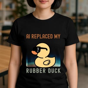 May include: Black t-shirt with the text "AI REPLACED MY RUBBER DUCK". The shirt features a cartoon yellow rubber duck wearing sunglasses. The background has a blue and green gradient.