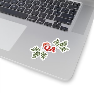 Op de afbeelding: Een witte sticker met de rode letters "QA", een kerstmuts en groene takjes. De sticker bevindt zich op een laptop. Het ontwerp suggereert een feestelijk kerstthema, perfect voor seizoensdecoraties.