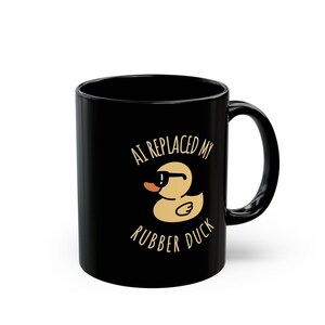 May include: Black ceramic mug with a humorous design. The mug features the text "AI REPLACED MY RUBBER DUCK" around a cartoon duck wearing sunglasses. The duck is yellow with a brown beak and a white belly.