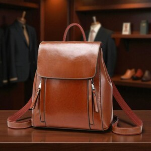 K&ouml;nnte beinhalten: Ein brauner Lederrucksack mit Klappenverschluss und zwei Rei&szlig;verschlusstaschen vorne. Die Tasche hat einen oberen Griff und verstellbare Schultergurte. Der Rucksack besteht aus gl&auml;nzendem, glattem Leder.