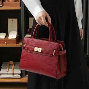 May include: A red handbag with a crocodile skin effect. The bag features two top handles and a gold-coloured clasp. The bag is held by a person wearing a white shirt and black dress.