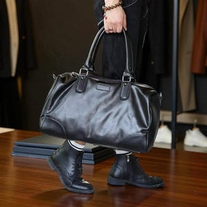 May include: A black leather duffel bag with two top handles and a shoulder strap. The bag has a rectangular shape with rounded corners and a small rectangular patch on the front. The bag is being held by a person wearing black boots.