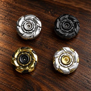 May include: Four circular metal fidget spinners arranged in a square formation. One is silver, one is matte black, one is gold with black accents, and one is a combination of silver and gold. Each features a central bearing and gear-like edges.