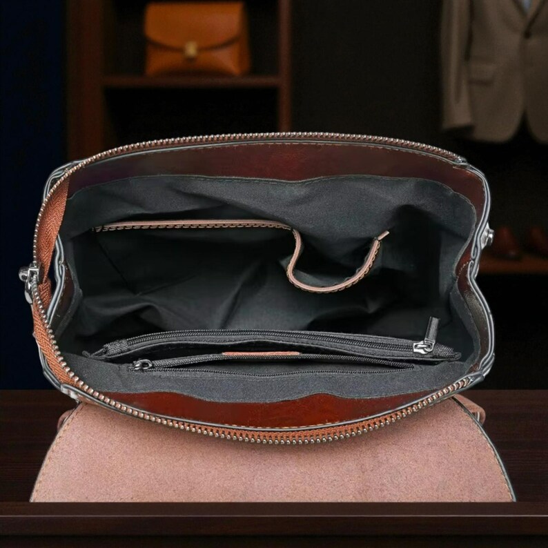 K&ouml;nnte beinhalten: Offene Ansicht eines braunen Lederrucksacks mit Rei&szlig;verschluss oben und Klappenverschluss. Der Innenraum ist schwarz mit mehreren F&auml;chern und einer kleinen Rei&szlig;verschlusstasche. Die Tasche hat einen braunen Riemen und silberne Hardware.