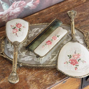May include: A vanity set featuring an oval metal tray with an ornate filigree border, a hand mirror, a hairbrush, and a comb. Each piece has an off-white surface decorated with pink rose floral motifs and intricate gold-toned metal handles.
