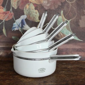 May include: A stack of white enamelware pots and pans with silver-colored handles. The set includes various sizes of pots and pans, with a small logo on the side of the largest pot. The background features a floral pattern.