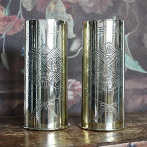 May include: A pair of polished brass cylindrical vessels featuring engraved floral motifs and a central emblem with the text ROYAL. The metallic surfaces are reflective, showing detailed line work of stems and leaves surrounding the crest.