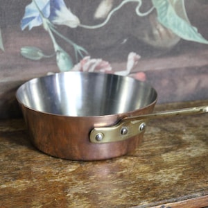 May include: A small copper saucepan with a brass handle. The interior is stainless steel. The pan is on a wooden surface. The handle is attached with rivets.
