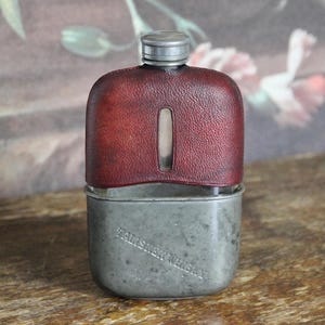 Pode incluir: Um frasco vintage com uma capa de couro vermelho e uma base de metal. O frasco tem uma pequena janela retangular e uma tampa de rosca de metal. As palavras "TALISKER WHISKY" estão gravadas na base de metal.
