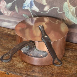 May include: A copper cooking pot with a lid and two dark metal handles. The pot is round and has a shiny surface. The handles are riveted to the pot. The pot is on a wooden surface.