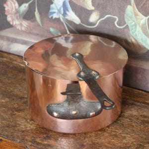 Può includere: Una pentola di rame con coperchio e manico in metallo scuro. La pentola ha una forma cilindrica e poggia su una superficie di legno. La pentola ha un manico e rivetti in metallo scuro. Lo sfondo presenta un dipinto floreale.