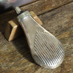 May include: An antique silver flask with a ribbed pattern on the lower section. The flask features a small spout and a metal cap. It is displayed on a wooden stand, set against a wooden surface.