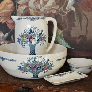 May include: A cream ceramic washbasin set comprising a jug, bowl, small bowl, and soap dish. Each item is adorned with a floral basket design in blue, purple, and green. The set is presented on a wooden surface.