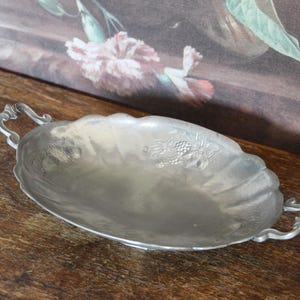 Könnte beinhalten: Ovale, silberfarbene Metallservierschale mit verzierten Griffen. Die Schale hat einen gewellten Rand und ein Blumendesign im Inneren. Die Schale ist wahrscheinlich Vintage und steht auf einer Holzoberfläche.