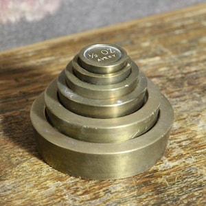 Puede incluir: Una pila de pesas antiguas de latón, dispuestas en tamaño descendente, con la pesa más pequeña en la parte superior. La pesa superior está etiquetada como "1/2 OZ AVERY". Las pesas son de color latón cálido y están sobre una superficie de madera.