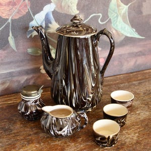 Può includere: Un set da caffè vintage con un motivo marmorizzato marrone e bianco. Il set comprende una caffettiera alta con coperchio, una lattiera, una zuccheriera e tre tazzine. Il set è esposto su una superficie di legno.