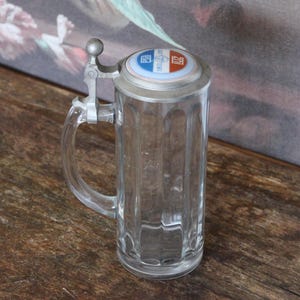 Peut inclure: Une chope à bière en verre transparent avec un couvercle à charnière en métal. Le couvercle présente un motif bleu, blanc et rouge. La chope a une anse et des rainures verticales.