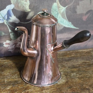 Può includere: Una caffettiera in rame con un manico in legno scuro. La caffettiera ha un lungo beccuccio e un coperchio con un piccolo pomello. La caffettiera è appoggiata su una superficie di legno.