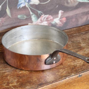 May include: A copper-clad saucepan with a silver interior and a dark metal handle. The pan is round and sits on a wooden surface. The handle is riveted to the side of the pan.