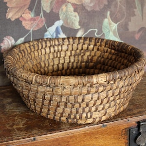 May include: A round, woven basket in natural light brown with darker brown detailing. The basket has a wide, open top and a textured surface. It is made from woven material, likely handmade. The basket rests on a wooden surface, suggesting a rustic aesthetic.