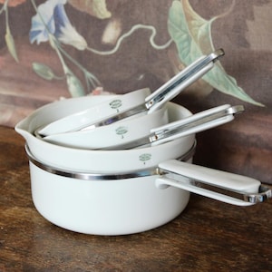 May include: A set of white enamel cookware with stainless steel handles. The set includes a large pot and three smaller saucepans nested inside. The handles are white with a silver trim. The cookware is arranged on a wooden surface.