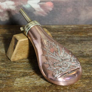 Könnte beinhalten: Eine kupferfarbene antike Pulverflasche mit detailliertem silberfarbenem Reliefdesign. Die Flasche hat einen Ausguss und ein dekoratives Relief von Kanonen und Flaggen. Die Flasche liegt auf einem Holzblock und zeigt ihre historische Ästhetik.