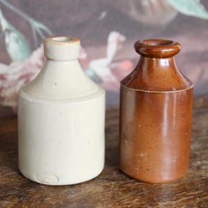 May include: Two antique stoneware bottles. One is off-white with a cylindrical body and a narrow neck. The other is brown with a similar shape. Both bottles have a rustic, aged appearance, suggesting they are vintage or antique items.