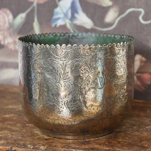 Puede incluir: Una maceta cilíndrica de metal con un borde superior festoneado y un acabado oscuro y deslustrado. La superficie exterior está cubierta con intrincados motivos florales y de aves en relieve, mientras que el interior muestra una pátina verde.
