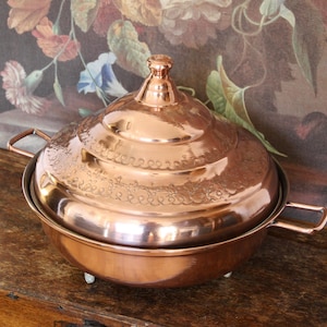May include: A copper serving dish with a lid, featuring a floral design. It has two handles and rests on small feet. The lid is topped with a knob. The dish is likely used for serving food, and is approximately 25cm in diameter.